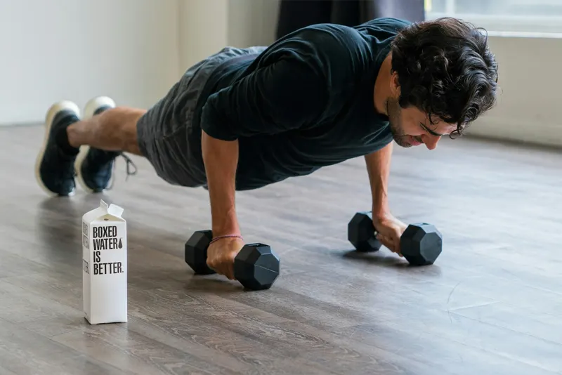 Aluno fazendo flexão com halteres no piso de madeira